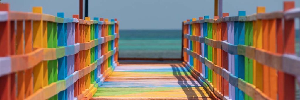 Solig brygga målad i regnbågens färger som leder ut mot ett blått hav.