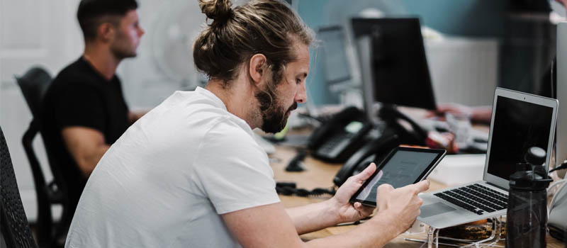 Man med vit t-shirt sitter och arbetar på sin ipad och laptop