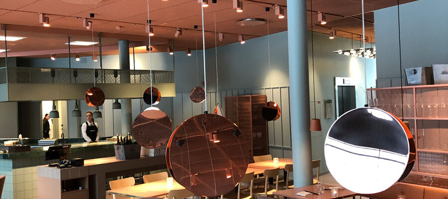 Modern restauranginteriör med kopparfärgade hängande lampor, öppet kök och personal i bakgrunden