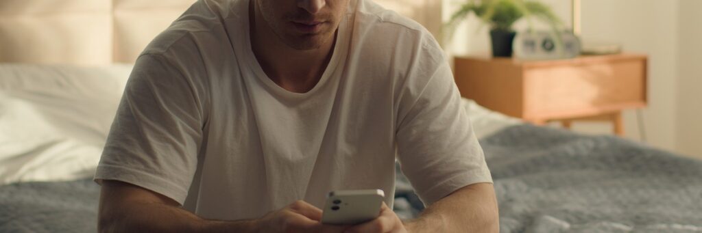 En man i vit t-shirt sitter på en säng och tittar på sin mobiltelefon