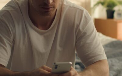 En man i vit t-shirt sitter på en säng och tittar på sin mobiltelefon