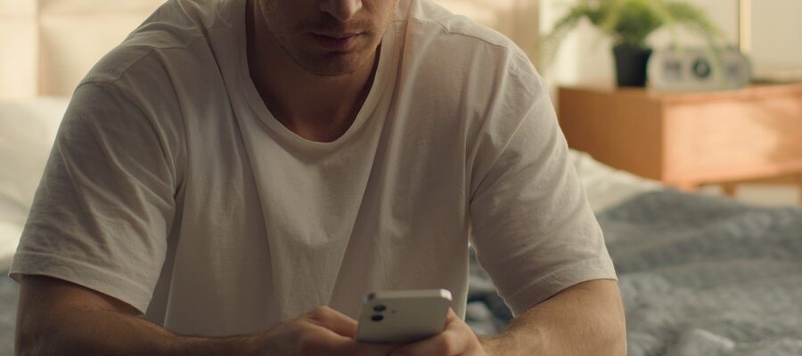 En man i vit t-shirt sitter på en säng och tittar på sin mobiltelefon