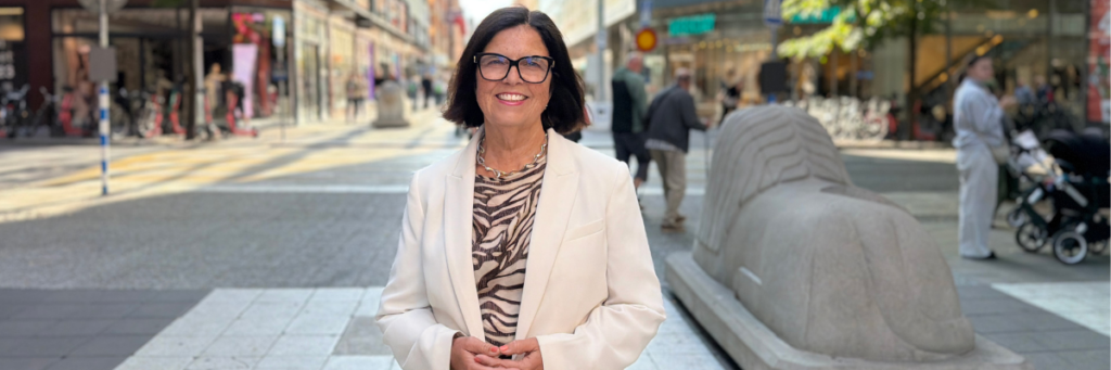 Leende kvinna i vit kavaj och zebramönstrad blus står utomhus på en gågata i centrala Stockholm.