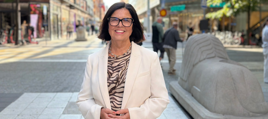 Leende kvinna i vit kavaj och zebramönstrad blus står utomhus på en gågata i centrala Stockholm.