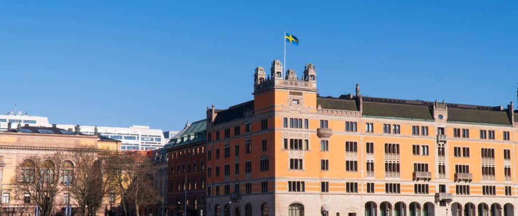 Historisk byggnad i orange fasad med svenska flaggan hissad på toppen mot klarblå himmel